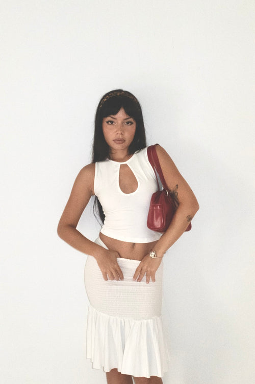 Woman wearing a white sleeveless top and skirt with a red handbag on a plain background